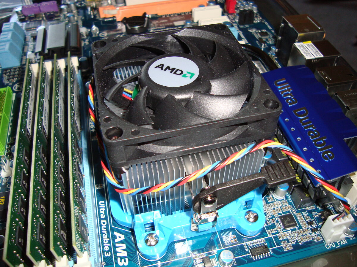 A black AMD fan and finned aluminum heatsink bolted to a blue motherboard, with four memory sticks standing to the left and striped fan wires running across the fins