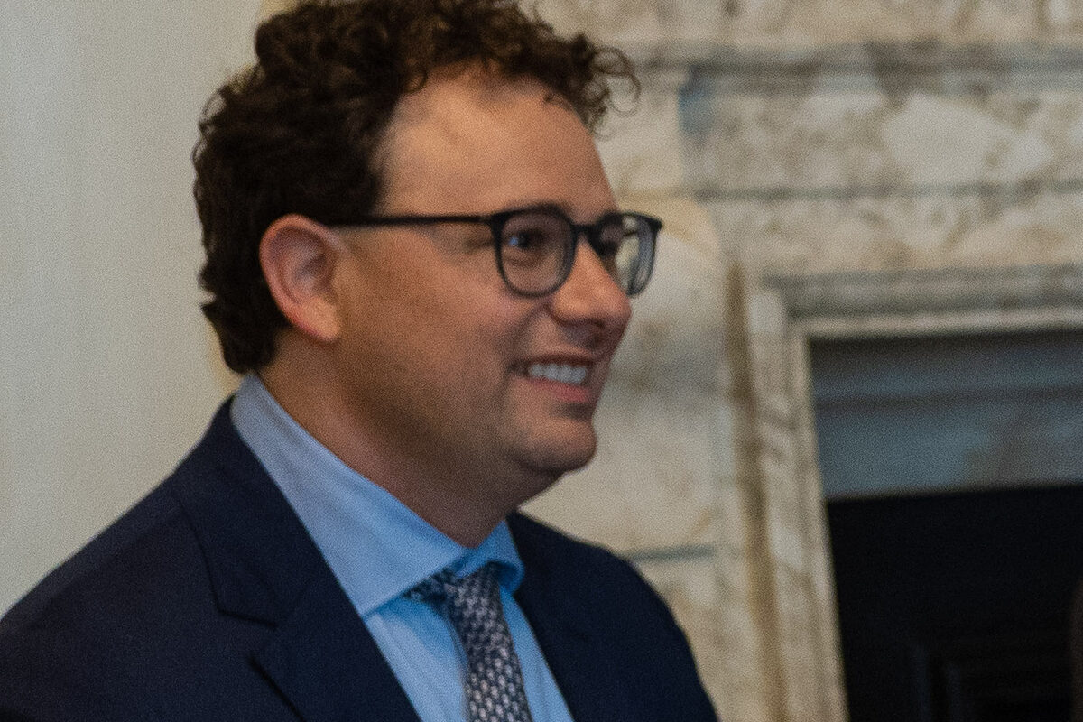 Dario Amodei, in glasses, a navy suit and a patterned tie, smiling in front of a marble fireplace.