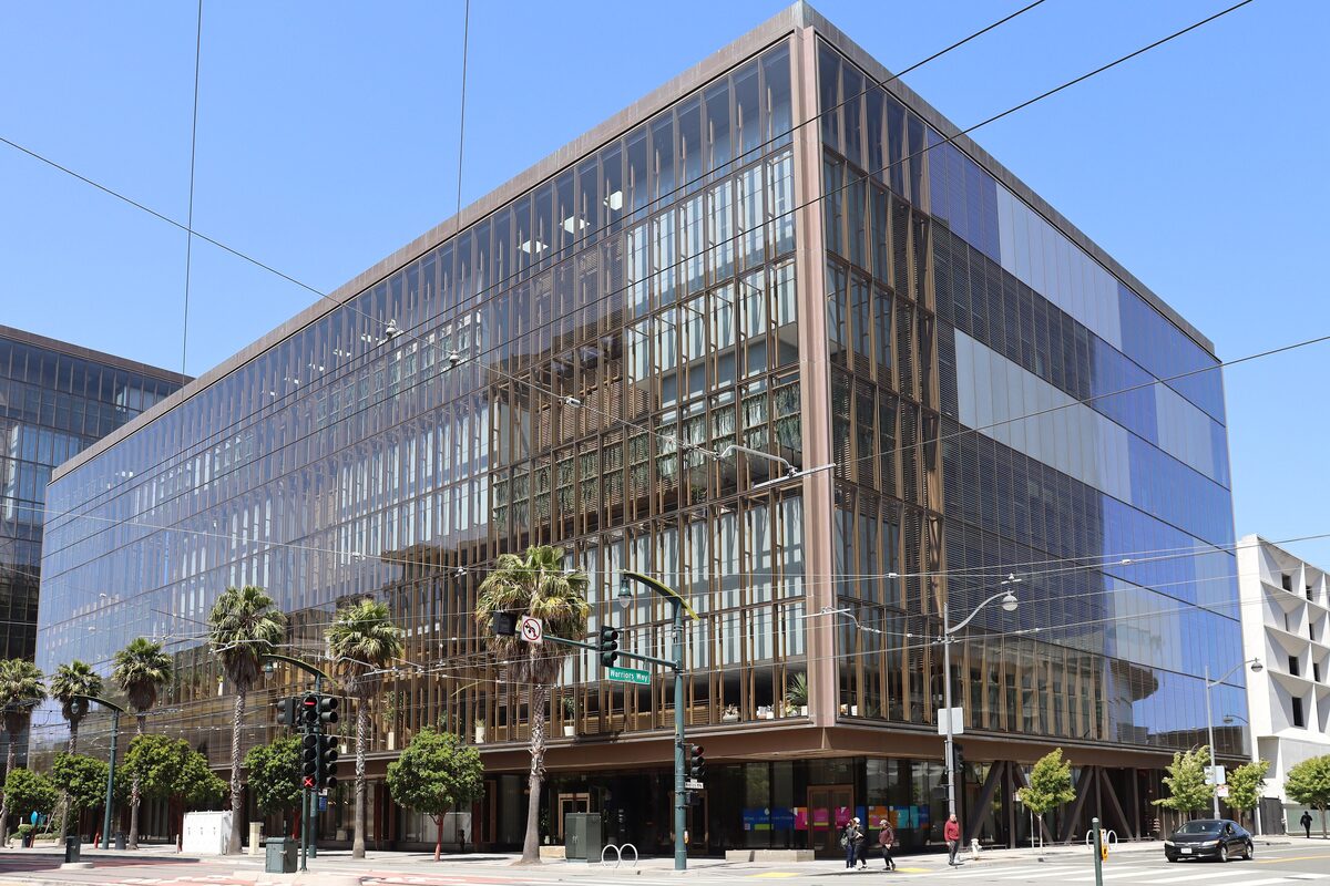 A large glass office building with a bronze-colored frame on a street corner, palm trees along the sidewalk under a clear blue sky.