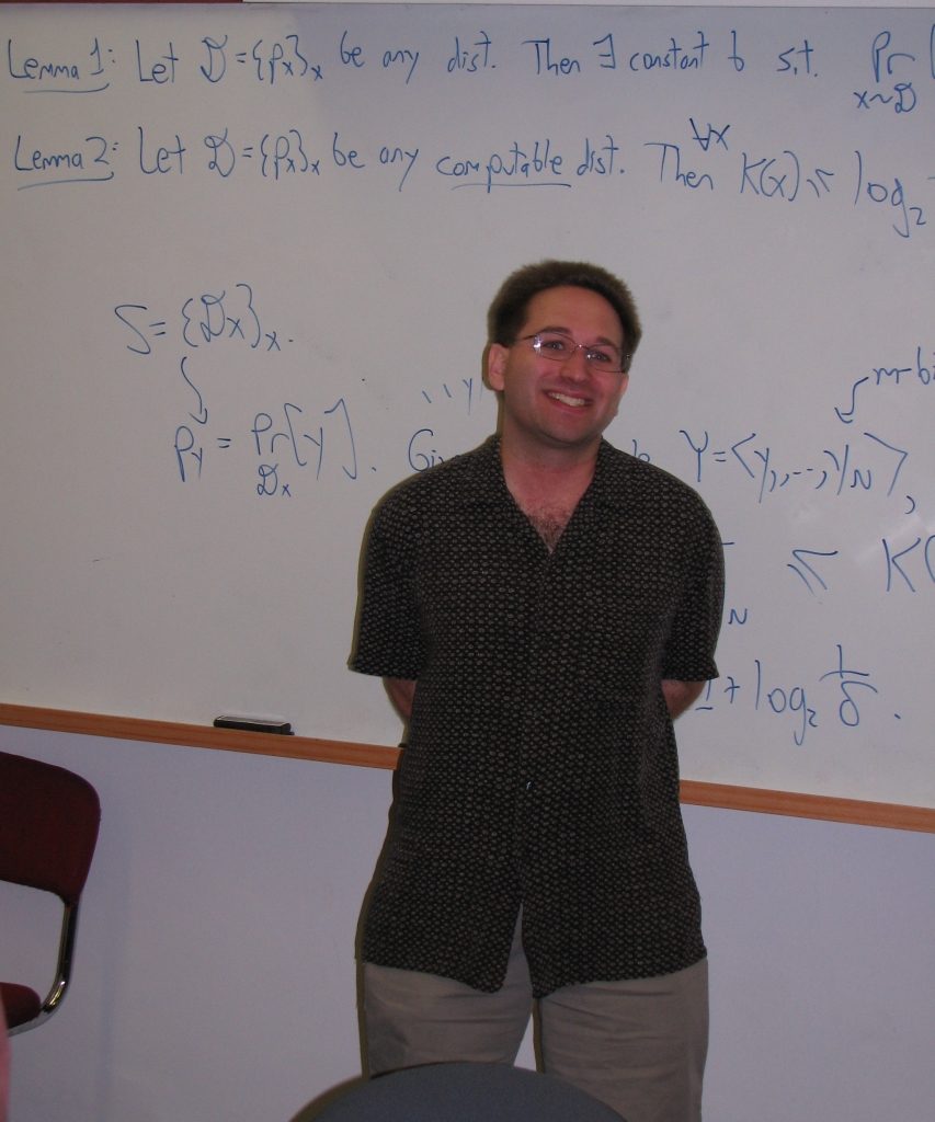 A smiling man in a dark patterned short-sleeved shirt standing in front of a whiteboard covered in handwritten lemmas and probability notation