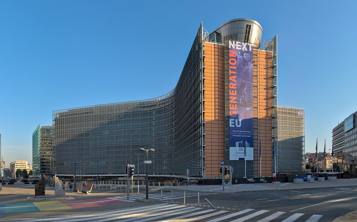 A long curved glass office building with a tall orange-paneled end section and a banner reading Next Generation EU, under a clear blue sky