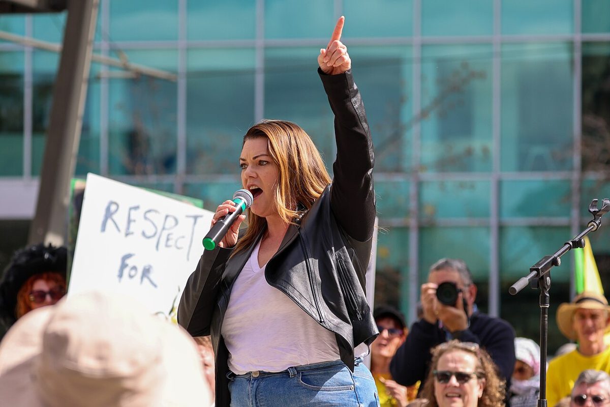 Sarah Hanson-Young in a black leather jacket, shouting into a microphone with one arm raised and finger pointing up, in front of a crowd and a glass building