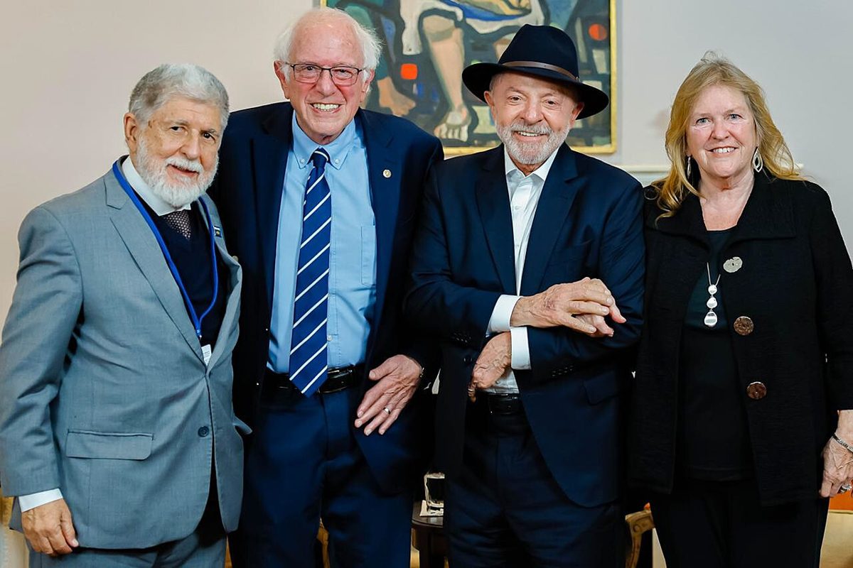 Four people smiling in front of a painting, with Bernie Sanders in glasses and a striped tie beside Lula in a dark fedora