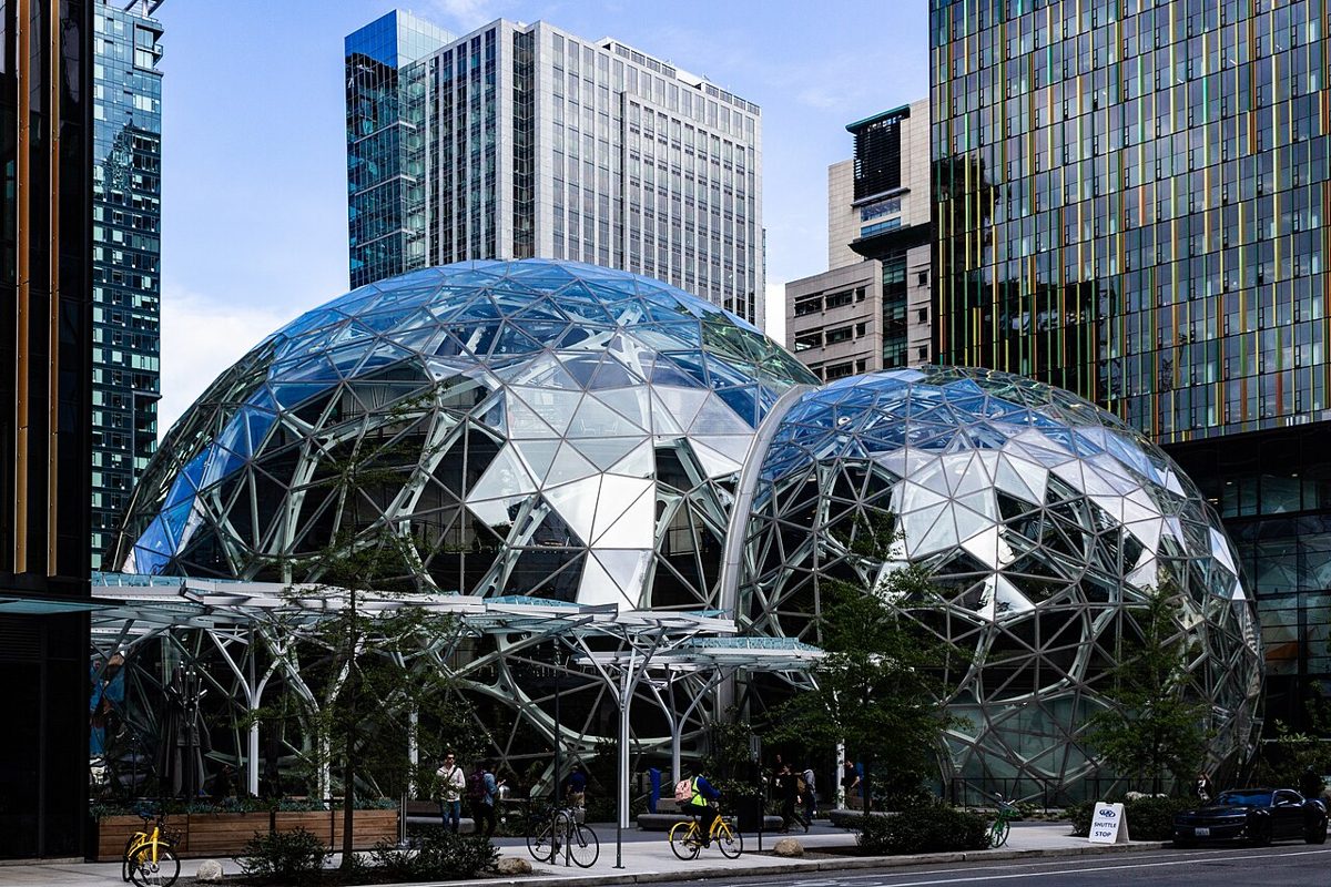 Two large glass domes with a lattice of steel triangles stand among office towers on a sunny street, with cyclists and pedestrians passing in front