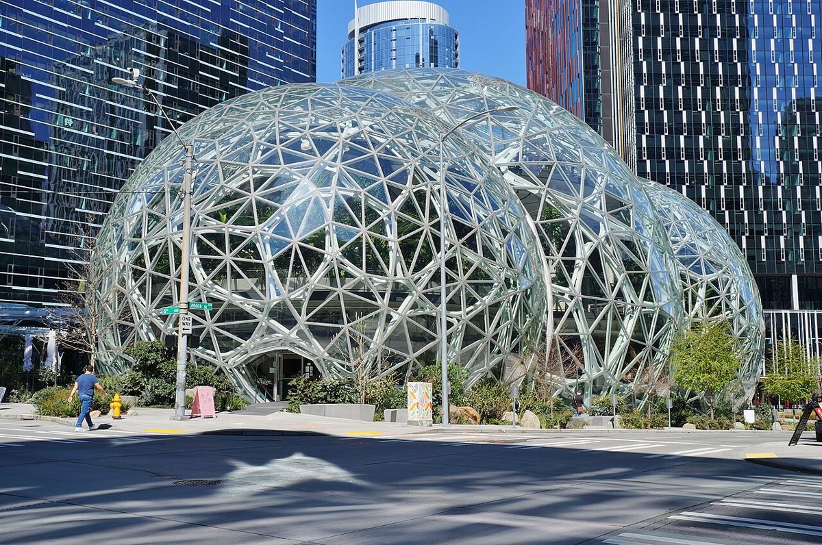 Three glass domes on a steel lattice frame at a street corner, with glass office towers behind them and a man walking past on the sidewalk