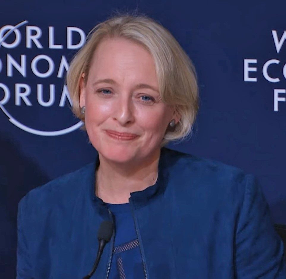 Julie Sweet in a blue jacket, smiling at a microphone in front of a dark blue World Economic Forum backdrop