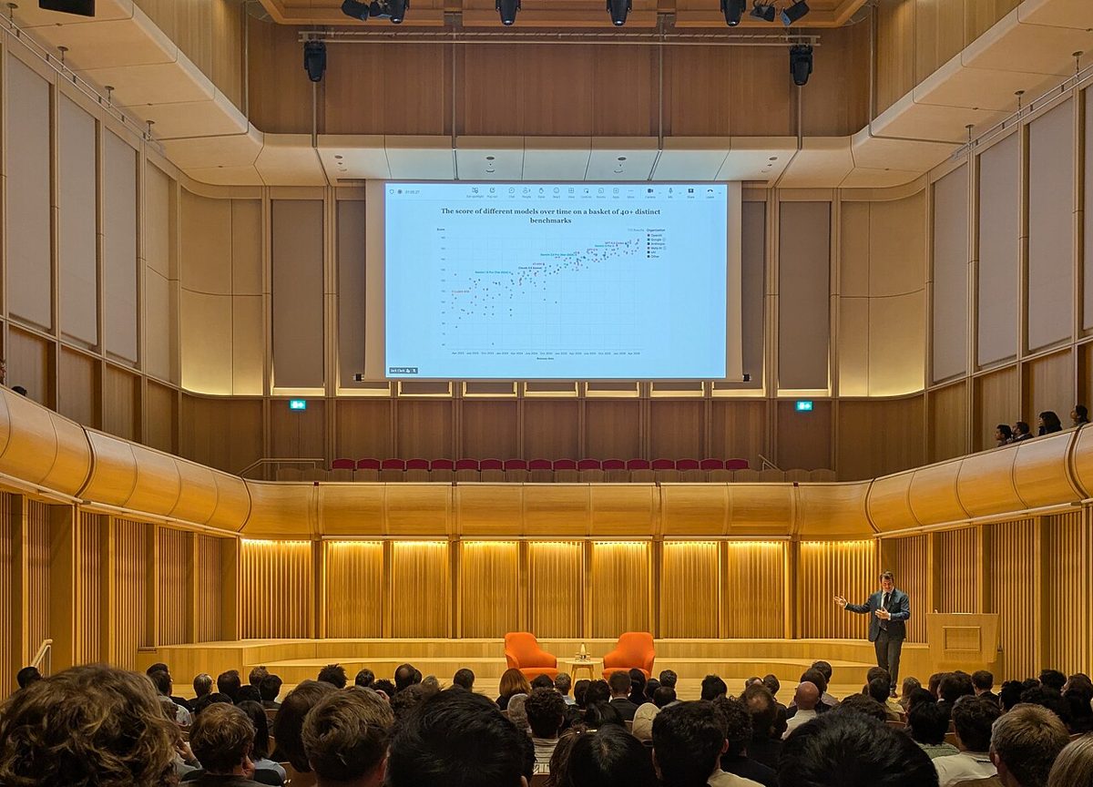 A packed wood-panelled auditorium seen from behind, a man in a suit speaking beside two orange armchairs, and a wide screen above him showing a scatter chart