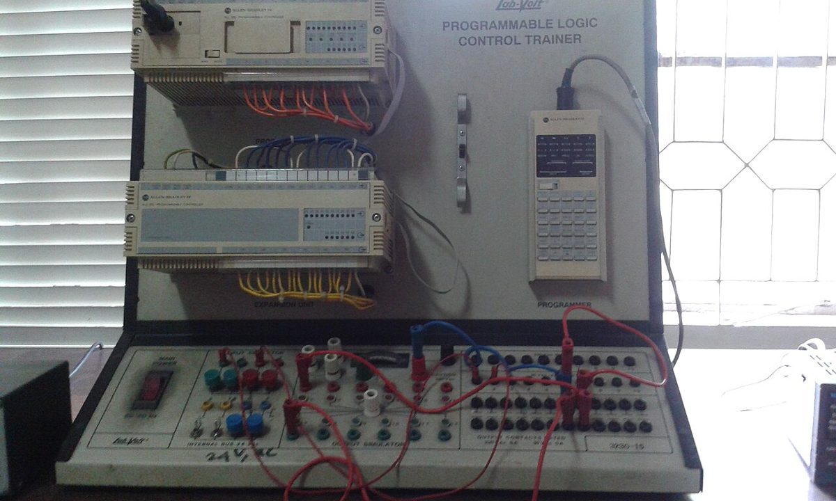 Two beige Allen-Bradley controllers wired with colored cables on a training bench, beside a handheld programmer and red patch leads