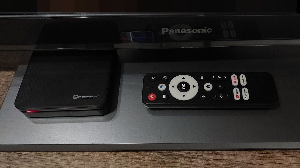 A small black Tracer set-top box with a red standby light, next to its remote control, on a gray stand below a Panasonic television