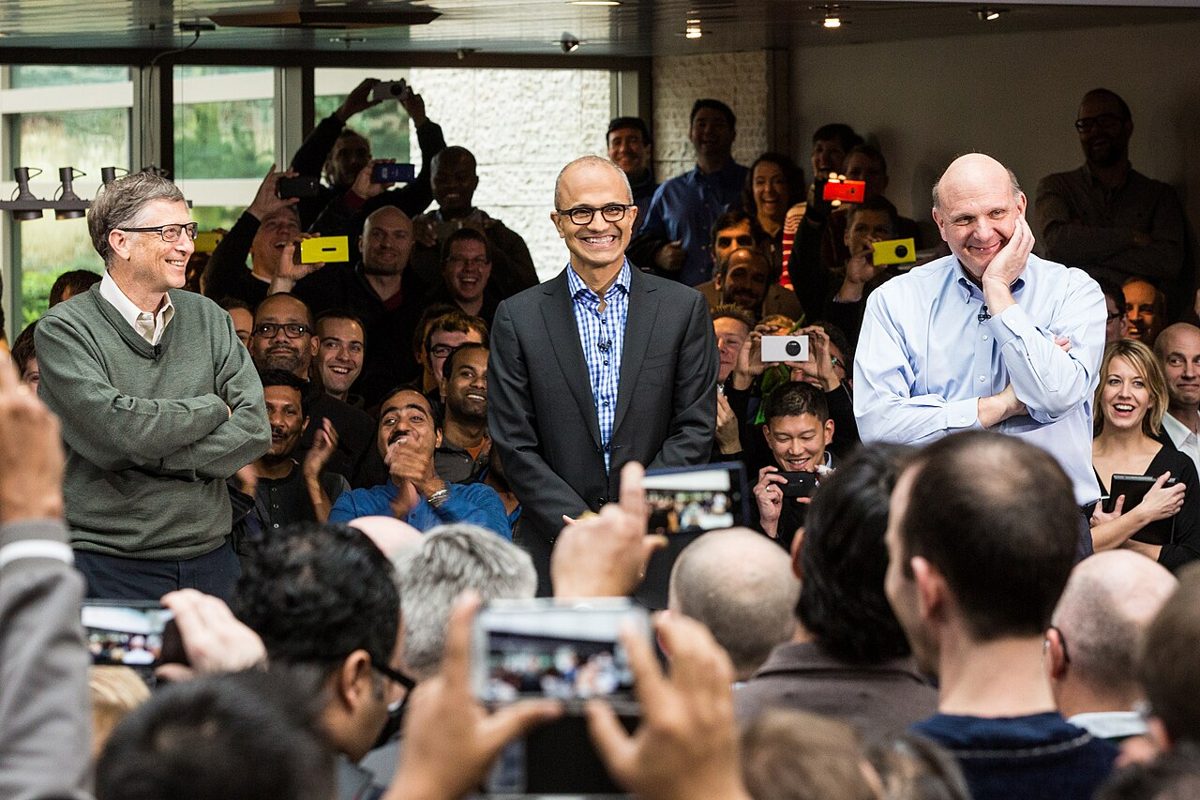 Bill Gates in a green sweater stands smiling with arms folded beside Satya Nadella and Steve Ballmer, before a crowd holding up phones