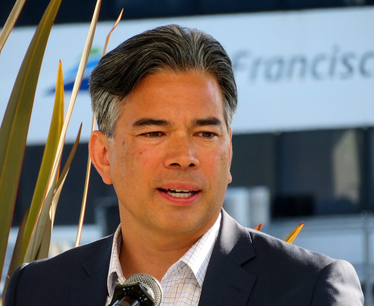Rob Bonta, gray-haired in a dark jacket and checked shirt, speaking into a microphone outdoors in front of a white ferry terminal building