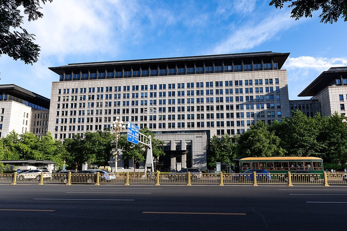 A wide, pale stone office block with rows of square windows and a dark overhanging roof, behind a gold-colored roadside railing on a broad, empty avenue