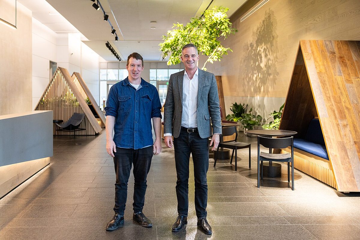Two men standing side by side in a bright office lobby with stone floors, a small indoor tree and a wooden alcove holding a blue bench