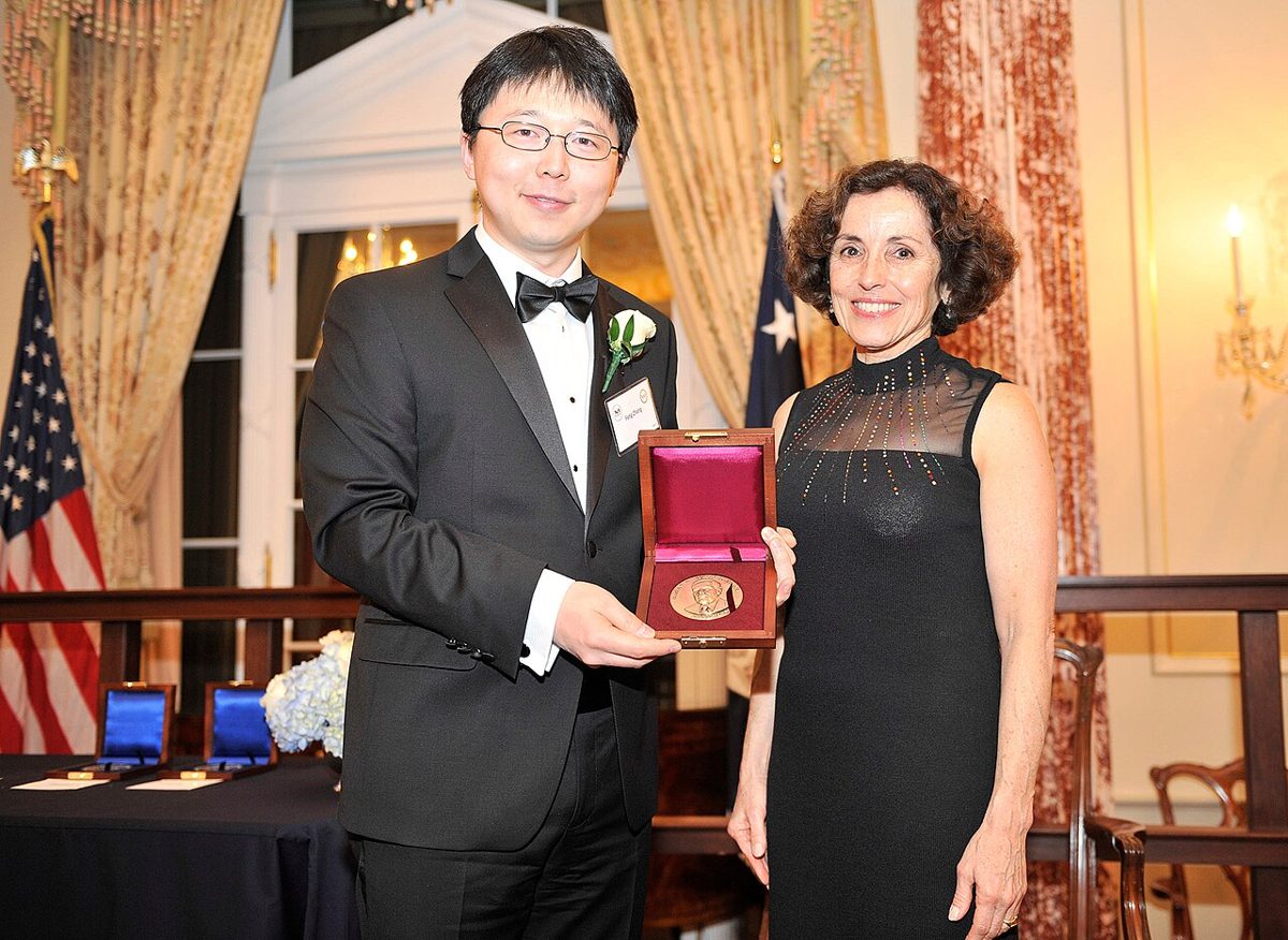 Feng Zhang in a tuxedo holds open a wooden case with a bronze medal beside a woman in a black dress, with American flags behind them