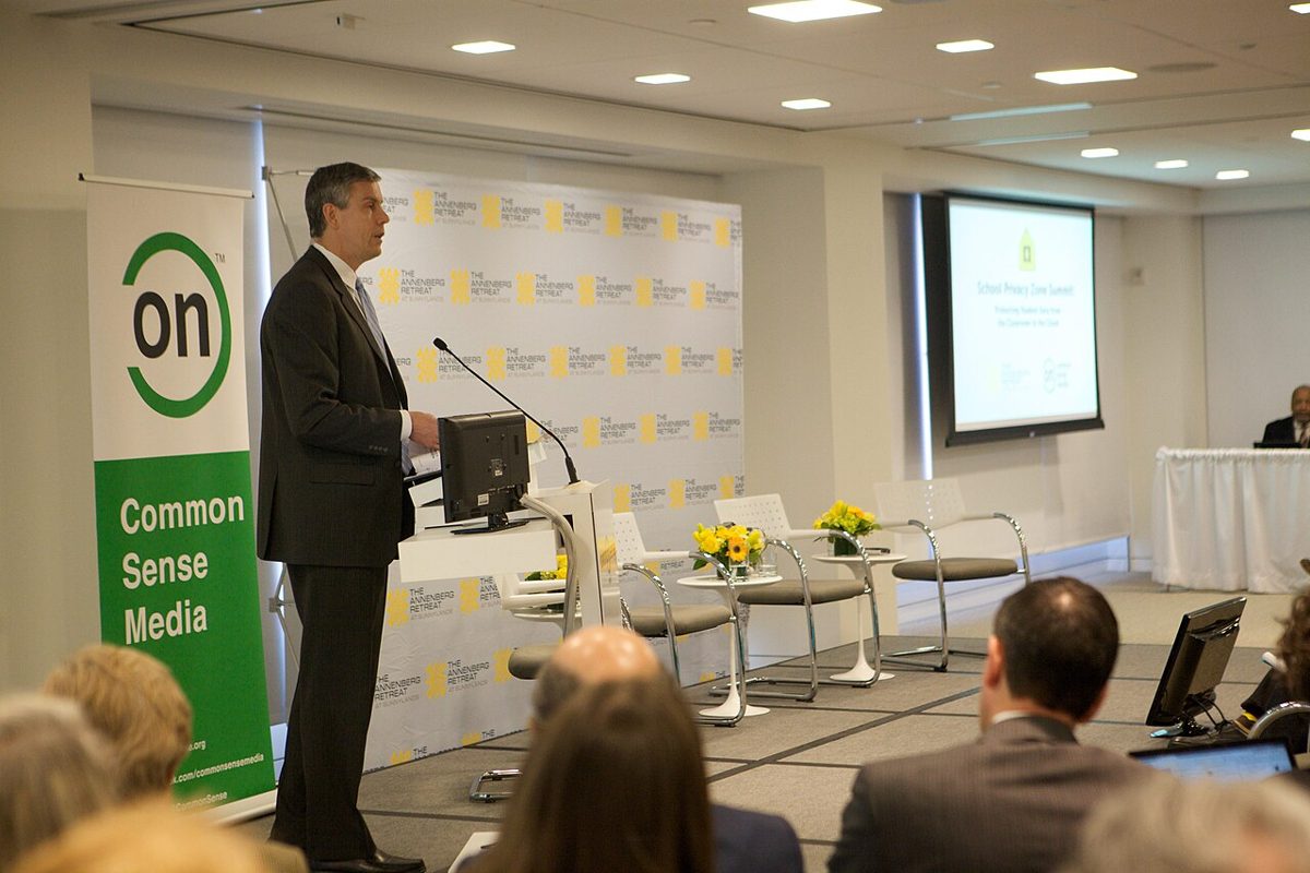 A man in a dark suit speaks at a podium beside a green Common Sense Media banner, facing a seated audience, with a presentation screen behind him