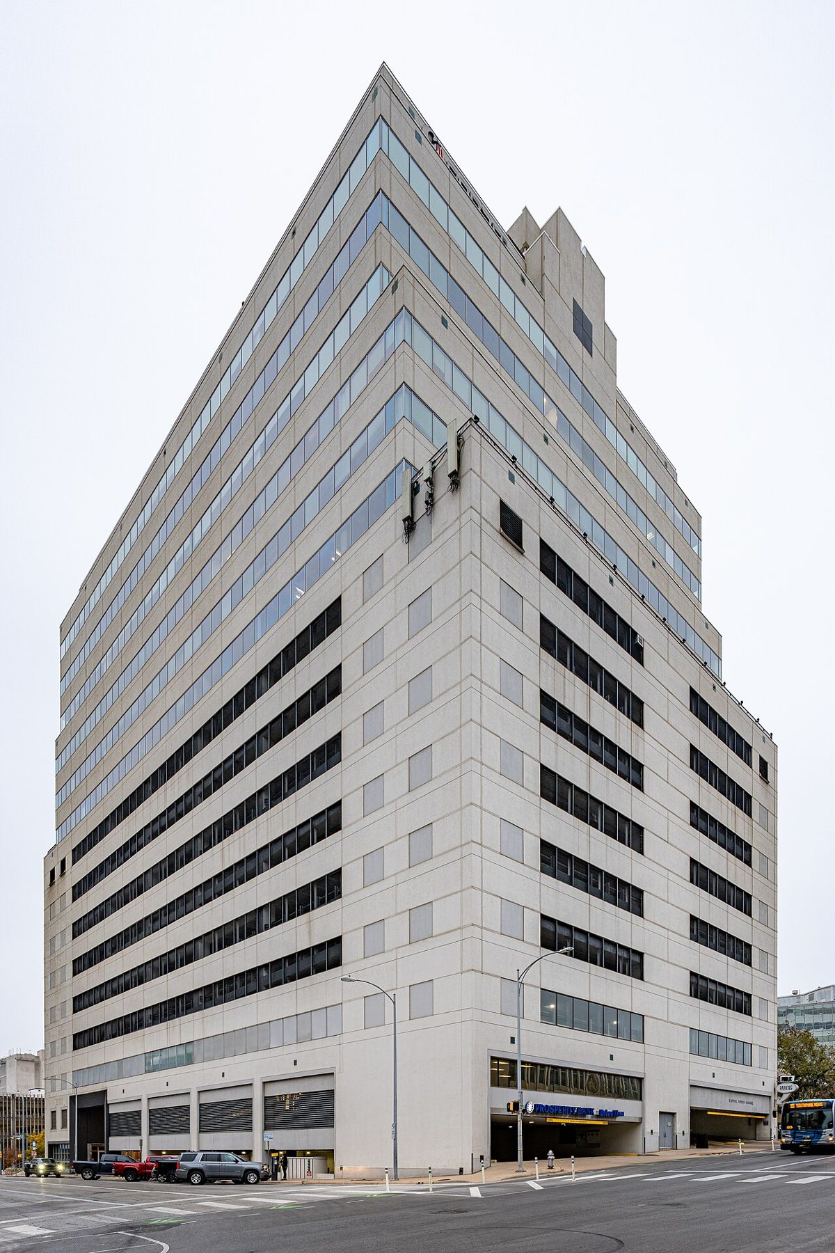 A tall pale concrete office tower with ribbon windows on a street corner under a white sky, cars parked along the curb and a city bus at the right