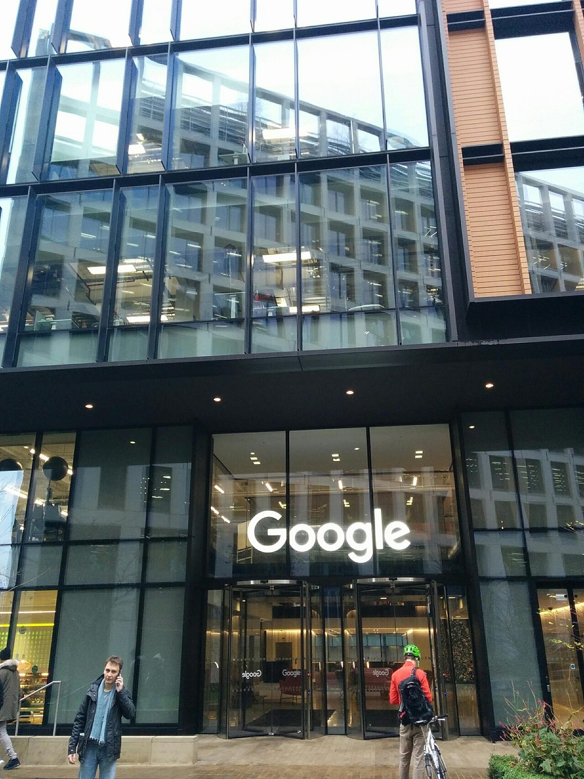 A glass-fronted office building with a lit Google sign above revolving doors, a man on a phone and a cyclist passing on the pavement outside