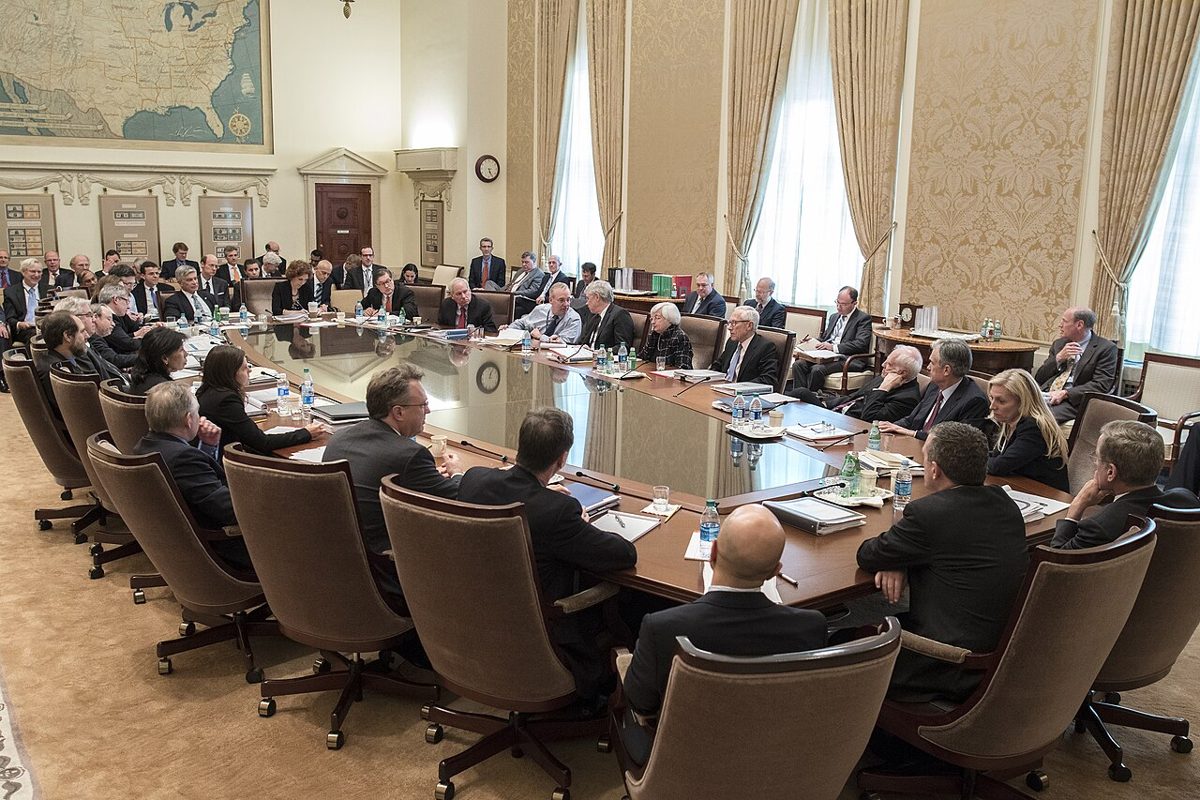 Officials in dark suits seated around a long polished table in a room with tall curtained windows and a large map of the United States