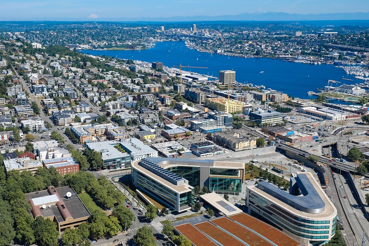 An aerial view of a curved white office complex in the foreground, with low city blocks, a blue lake full of moored boats and distant hills behind it