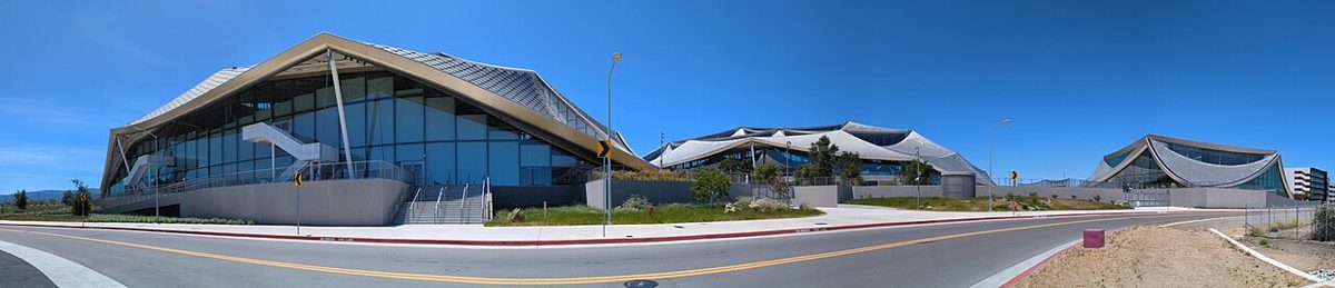 Three long buildings with curved, tent-like metal roofs and glass walls, set behind an empty road under a clear blue sky