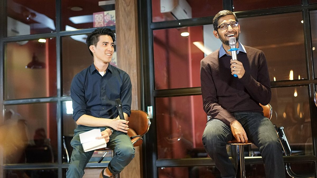 Sundar Pichai, in glasses and a dark sweater, smiles on a stool holding a microphone, beside an interviewer in a navy shirt with notes on his knee