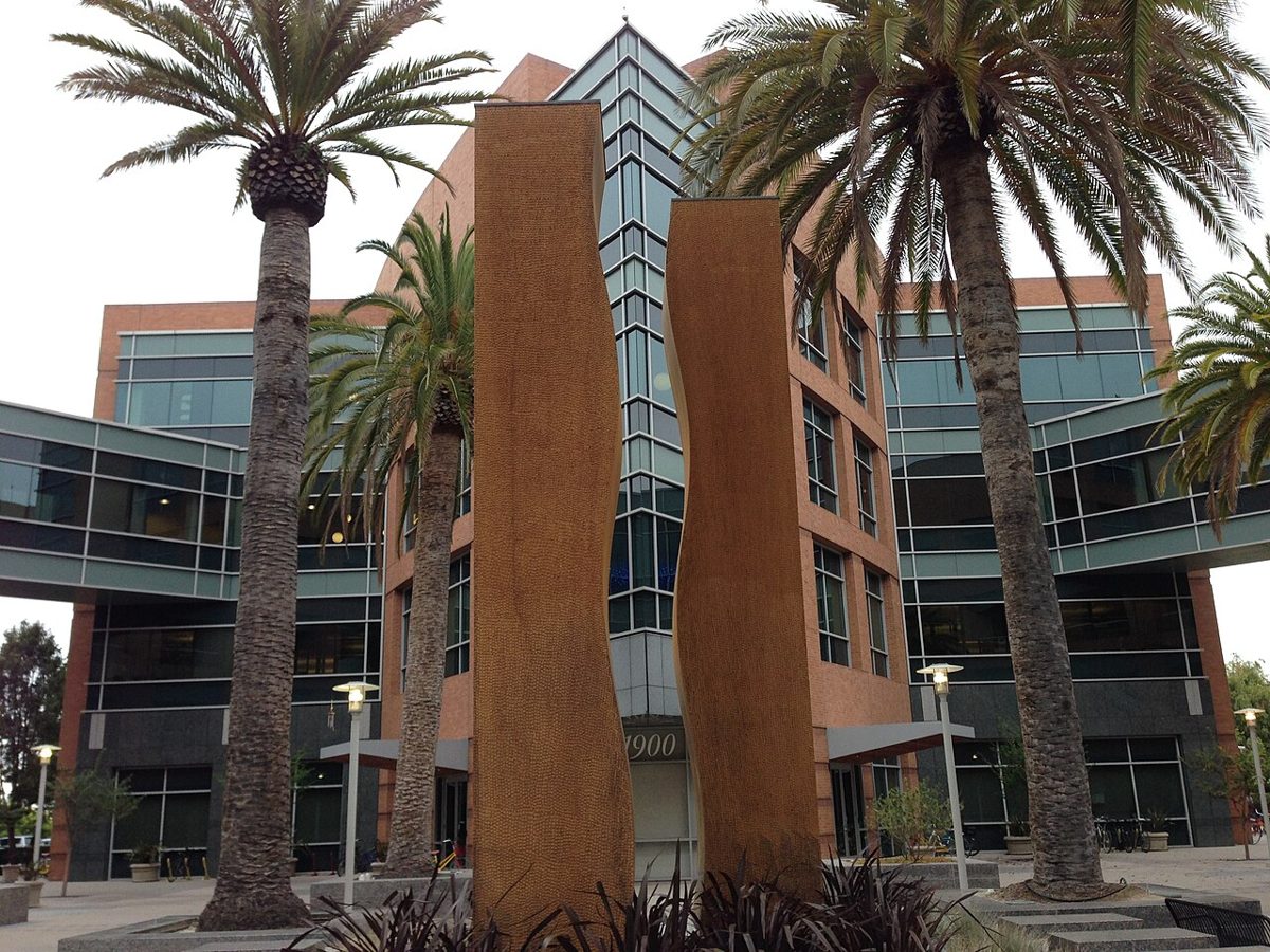 A red brick and glass office building numbered 1900, fronted by palm trees and two tall rust-colored sculptures with wavy edges