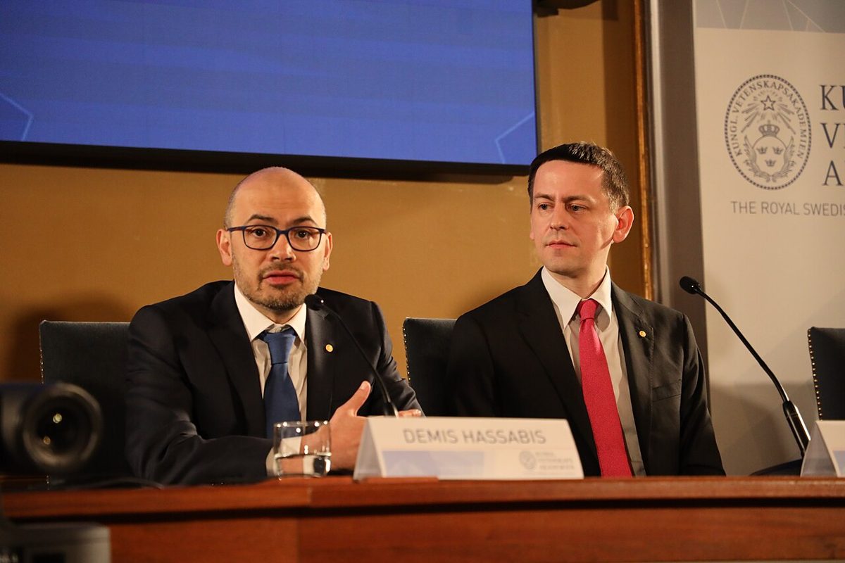 Demis Hassabis, in glasses, speaks at a wooden press table behind his name card, with John Jumper in a red tie beside him