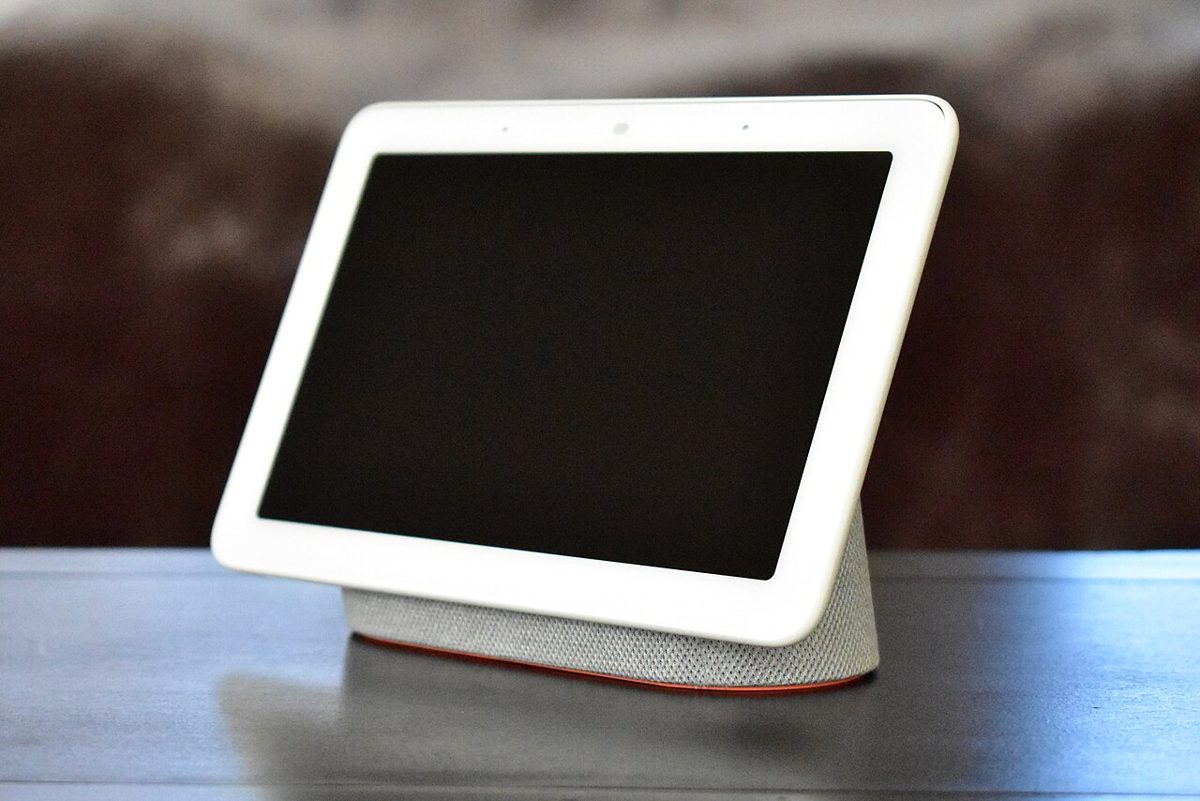 A white smart display with a blank screen and a grey fabric speaker base, sitting on a dark wooden table