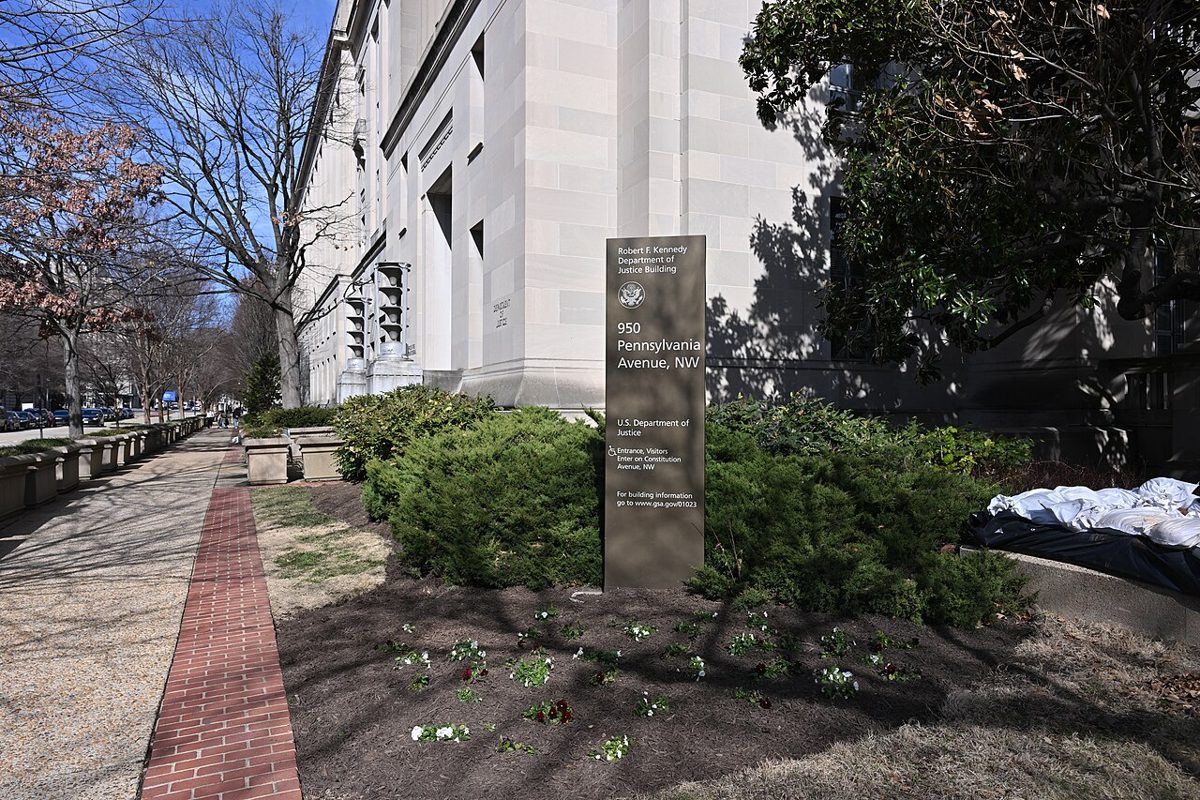 A pale stone government building on a tree-lined Washington street, with a brown sign in a flower bed reading Robert F. Kennedy Department of Justice Building, 950 Pennsylvania Avenue NW