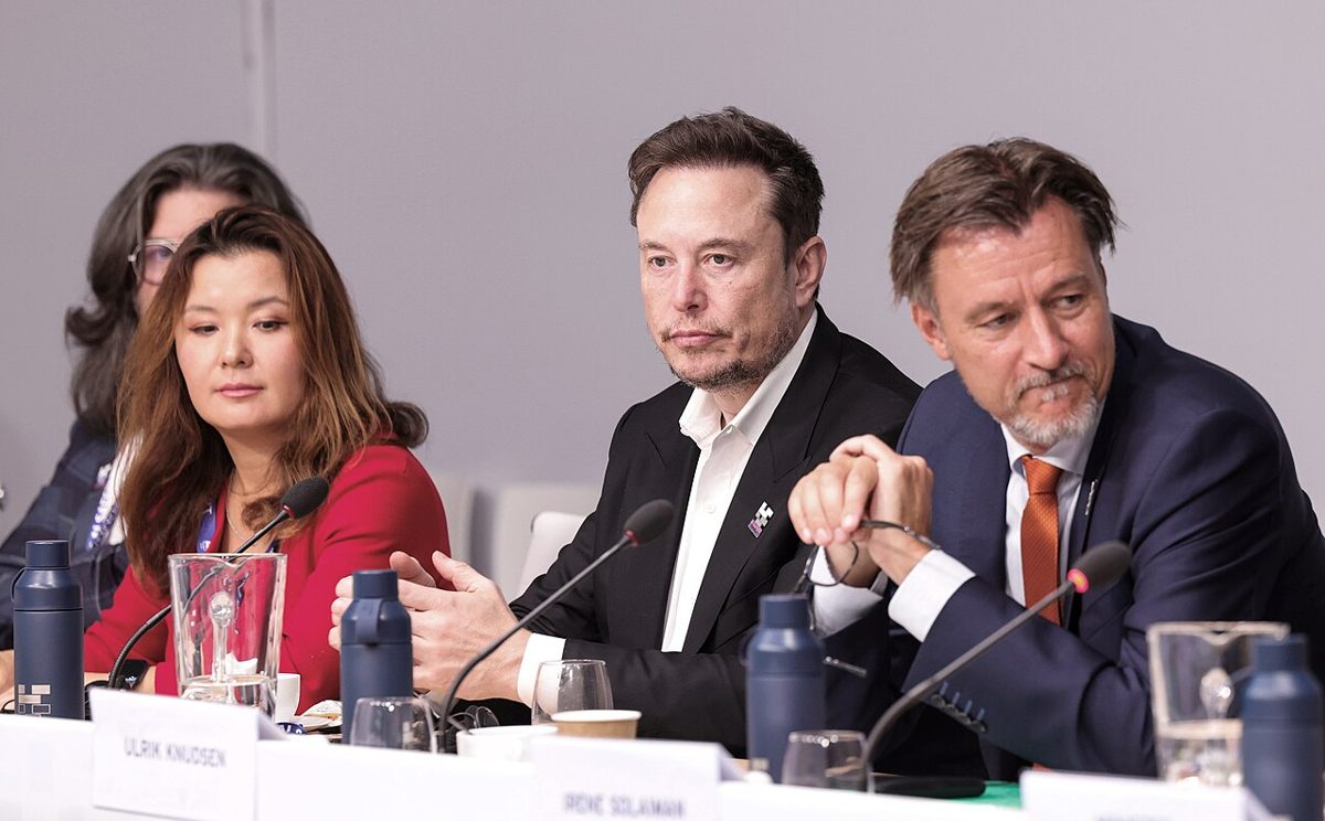 Elon Musk seated at a conference table with microphones and water bottles, between a woman in a red jacket and a man in a blue suit