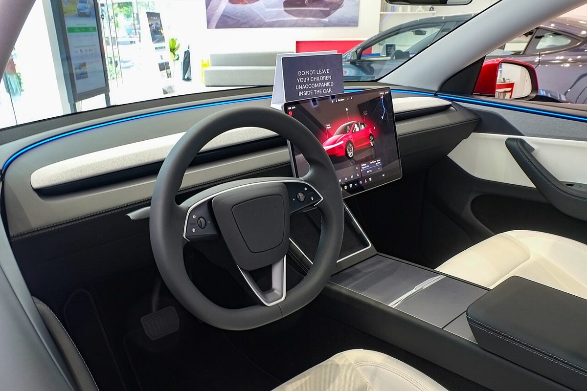 A Tesla cabin seen from the driver's door, with a steering wheel, a center touchscreen showing a red car, and a showroom sign about children on the dashboard