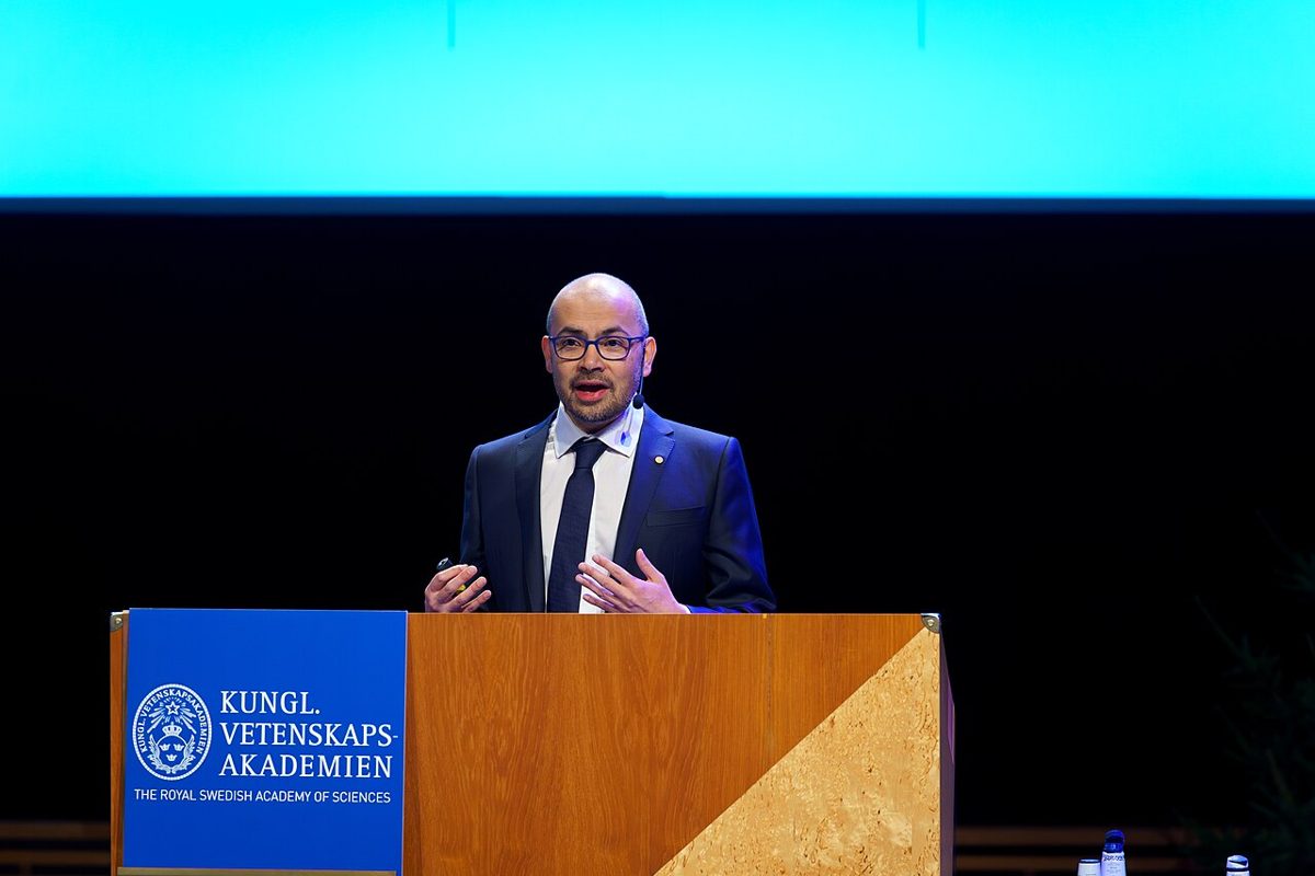 A man in a dark suit and glasses speaks from a wooden lectern marked Kungl. Vetenskapsakademien, lit blue behind him