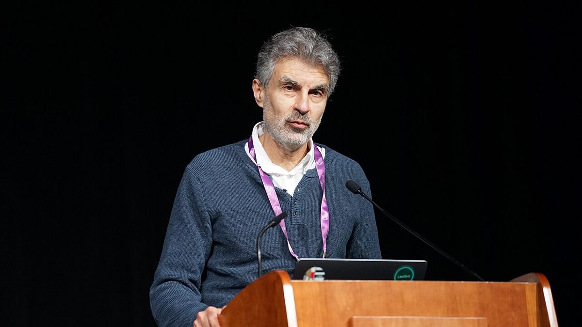 A gray-haired, bearded man in a blue sweater and purple lanyard speaks at a wooden lectern with a laptop against a black backdrop