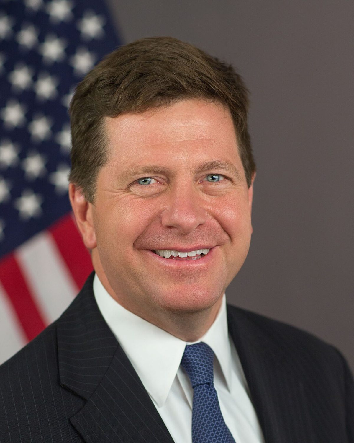 Jay Clayton in a dark pinstriped suit and blue tie, smiling, in front of an American flag