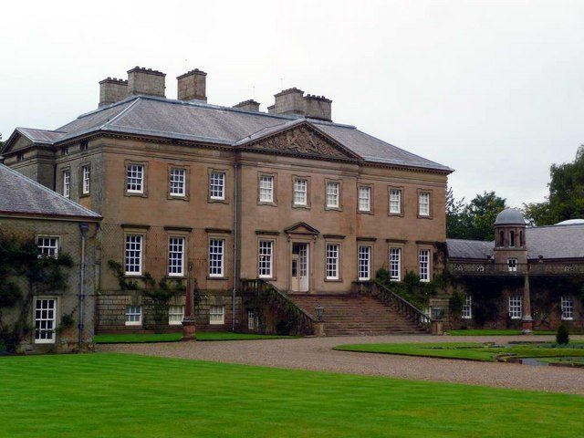 A grey stone Georgian country house with a carved pediment and twin curved stairs to the front door, across a mown lawn and gravel drive