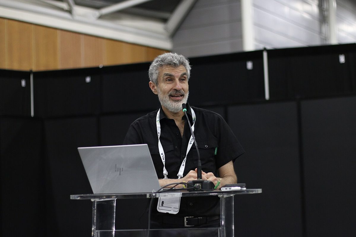 A gray-haired bearded man in a black shirt speaks into a microphone at a clear glass lectern with an open laptop on it, a conference lanyard around his neck