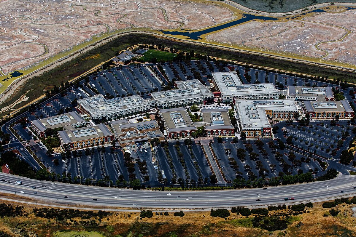 An aerial view of a cluster of low office buildings surrounded by parking lots, with a highway in front and salt marsh behind
