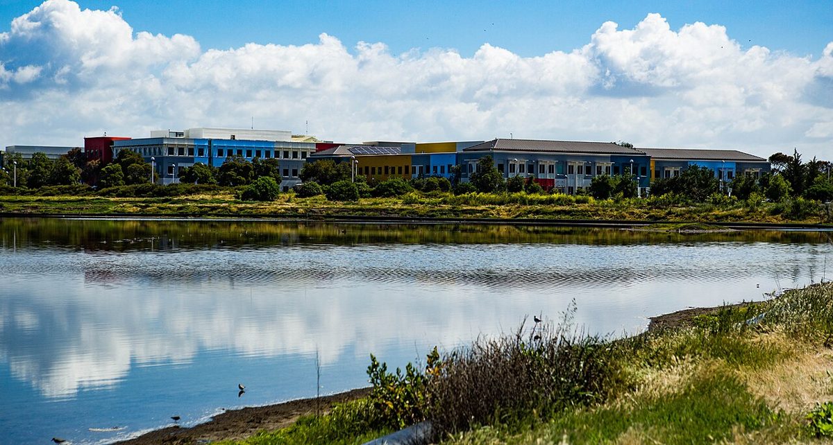 A row of low office buildings in blue, yellow and red behind trees, reflected in a still lagoon under white clouds