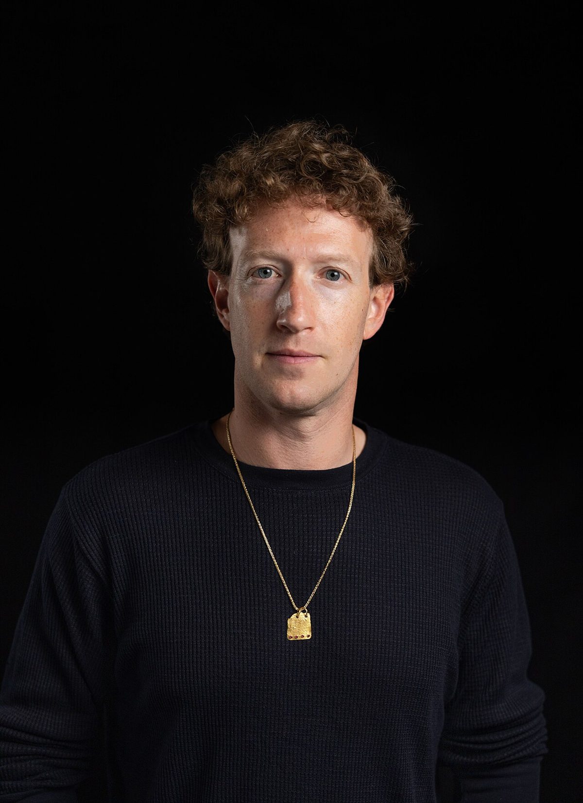 Mark Zuckerberg in a black sweater and a thin gold chain, looking at the camera against a plain black background