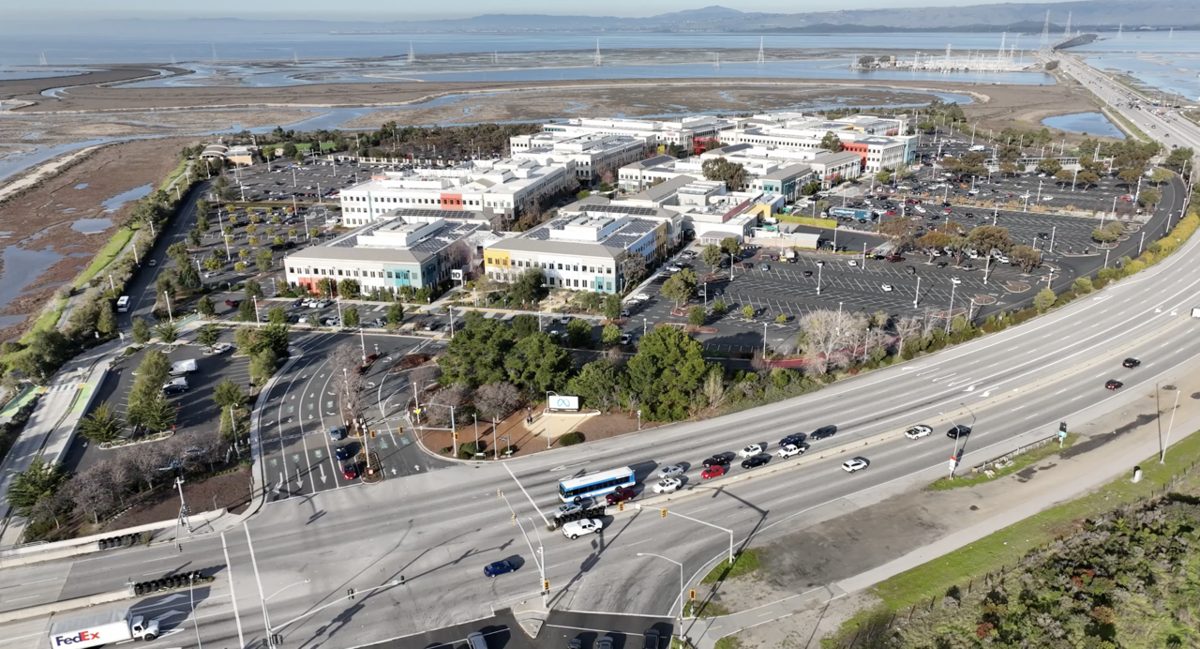 An aerial view of a low-rise office campus beside salt marshes, with large parking lots, a wide curving highway in front and the bay behind