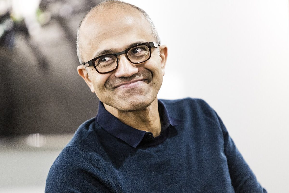 Satya Nadella in dark-rimmed glasses and a navy sweater over a collared shirt, smiling and looking off to one side against a pale background