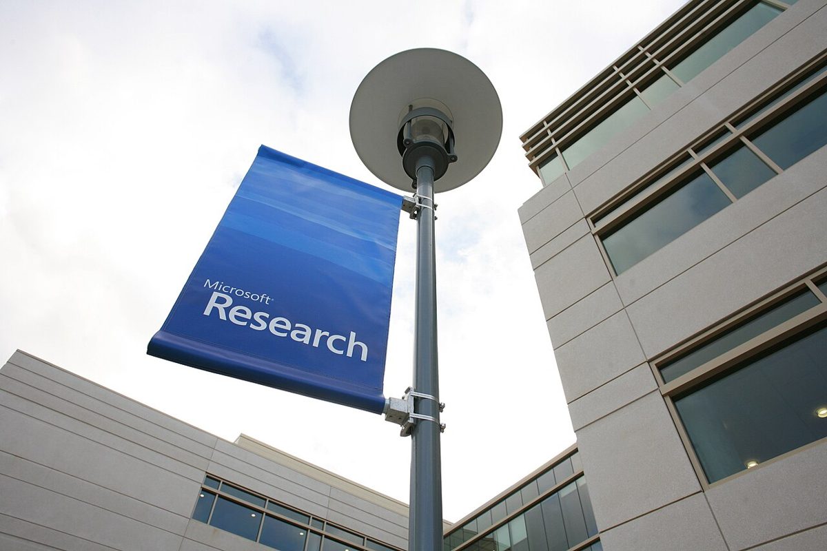 A blue Microsoft Research banner on a lamppost, seen from below, between two pale stone office buildings with long rows of windows