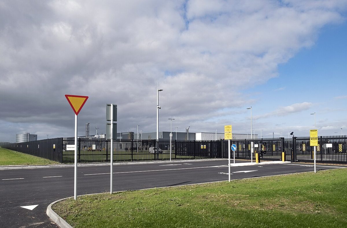 A long black security fence and gated entrance in front of low gray industrial buildings under a cloudy sky, with a yield sign and an empty road in the foreground