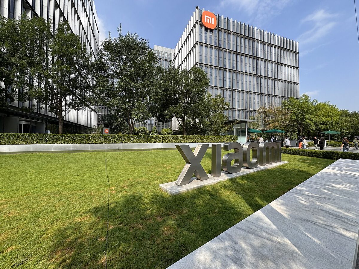 A brushed steel Xiaomi sign standing on a lawn in front of two glass office blocks, the taller one carrying the orange Mi logo above its top floor