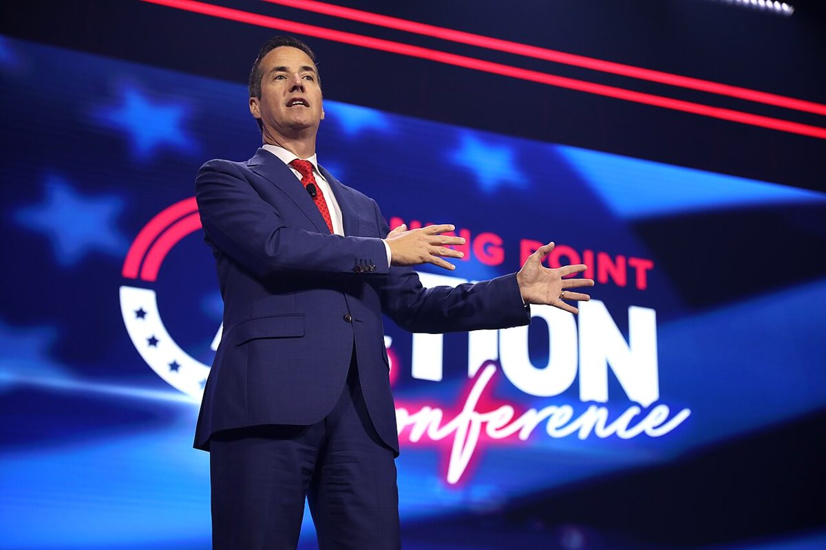 Bernie Moreno in a blue suit and red tie speaking on a stage with both hands out, in front of a large screen with stars
