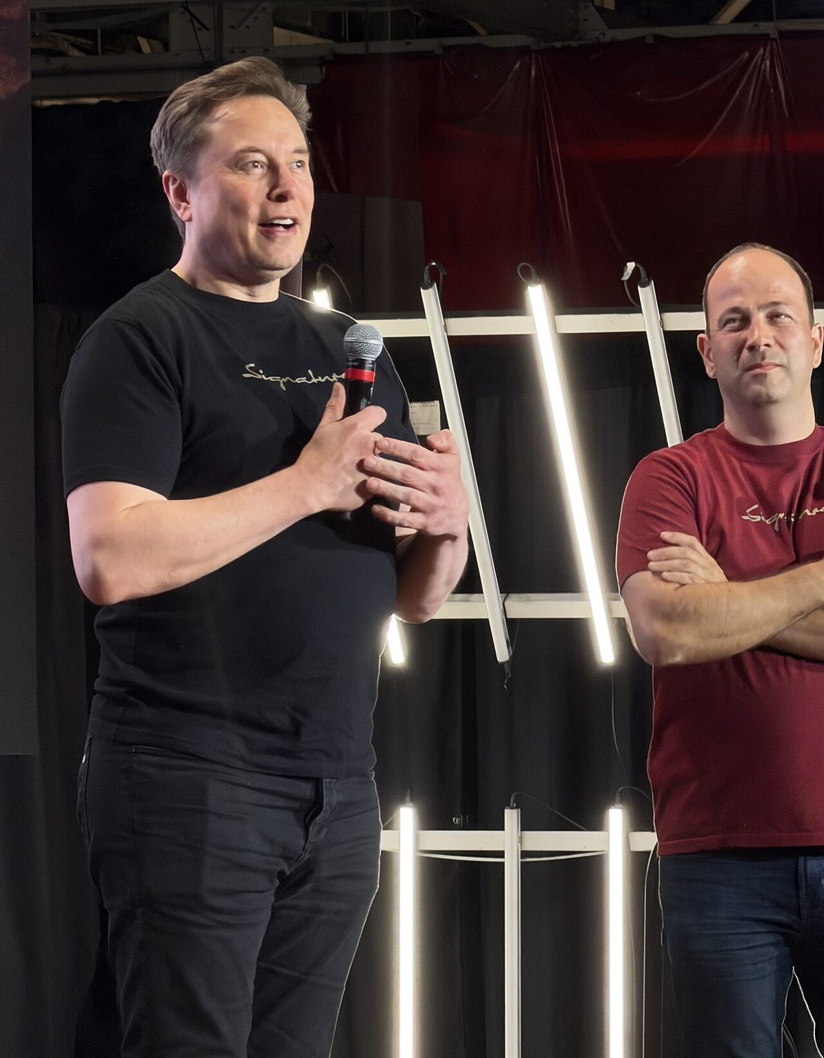 Elon Musk in a black T-shirt talking into a microphone on a stage lit by white tubes, beside a man with folded arms
