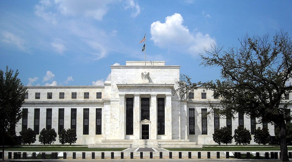 The white marble facade of the Federal Reserve Board building behind trees, with an American flag flying from the roof