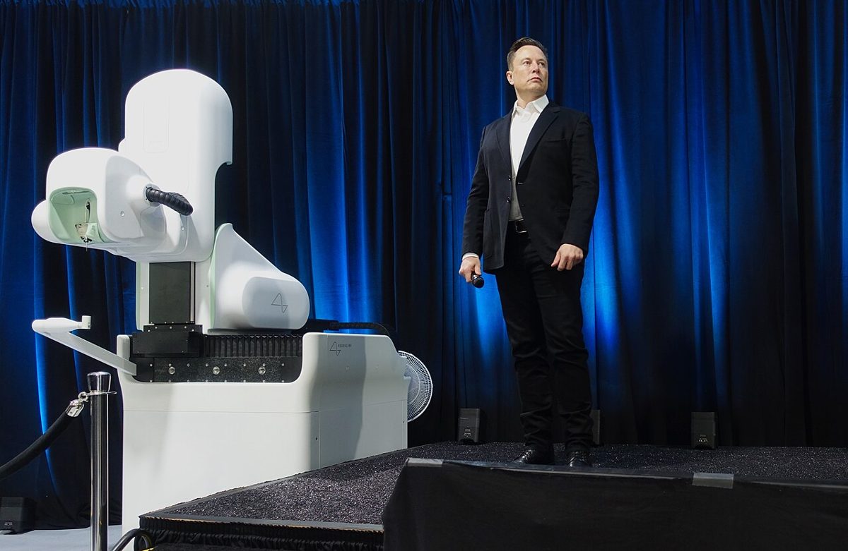 A man in a dark suit and white shirt stands on a stage with a microphone in hand, beside a white robotic machine, in front of a blue curtain