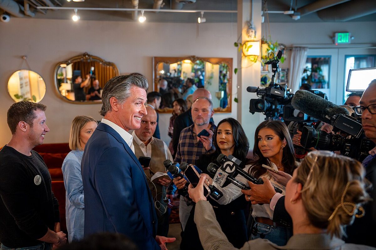 Gavin Newsom in a navy blazer smiling as reporters hold microphones and phones toward him, with TV cameras behind them