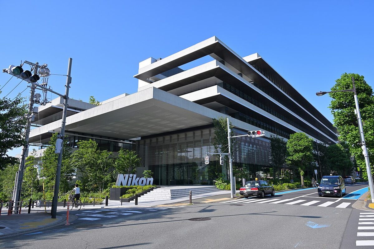 A white, layered office building with deep overhanging floors behind trees, a Nikon sign by the entrance steps and taxis at a crossing